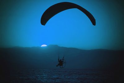 Person paragliding over sea by mountain at dusk