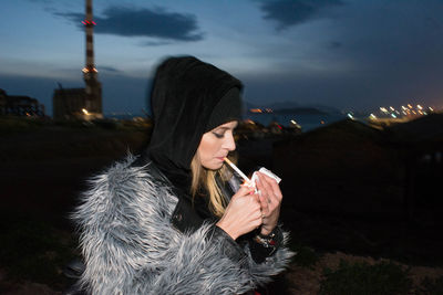 Young woman lighting cigarette in city against sky at night