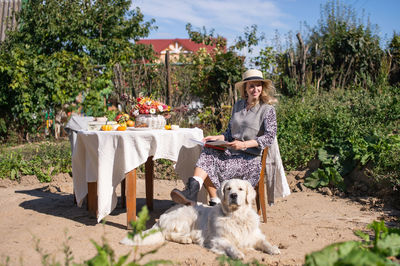 A woman in a hat is sitting on a chair near the table, a labrador retriever is lying next to her