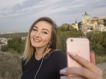 Woman listening music while taking selfie through mobile phone against sky