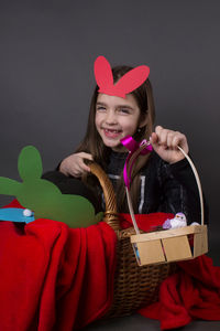 Portrait of smiling girl with costume rabbit ears sitting in basket against gray background