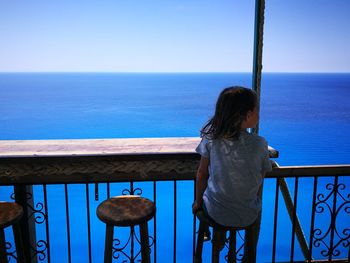 Rear view of girl sitting by railing against sea