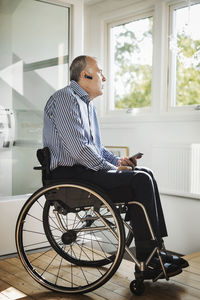 Mature man holding mobile phone while using bluetooth in wheelchair at home