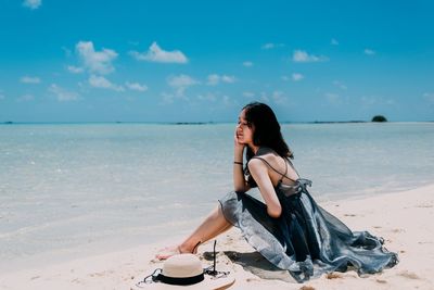 Side view of young woman sitting at beach against blue sky during sunny day