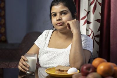 An indian asian woman holding a glass of milk sitting beside a table in a domestic room