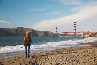 Rear view of woman standing on shore with golden gate bridge in background