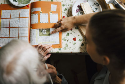 High angle view of female caregiver solving crossword puzzle with senior woman at table