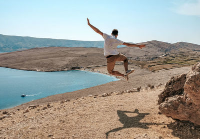 Rear view of young man jumping for joy on rocky coastline overlooking bay and beach.