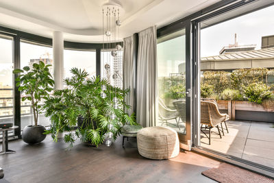 Fragment of interior design of recreation lounge area with green tropical plants and cozy poufs and stylish hanging lamp near panoramic window and terrace door in luxurious house