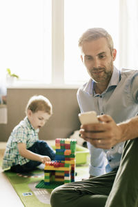 Smiling father using phone while son playing with toy blocks at home