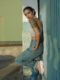 Confident young female with tattoos wearing stylish outfit leaning wall in city and looking at camera