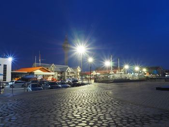 Illuminated street by buildings against sky at night