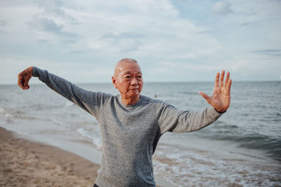 Senior man exercising while standing at beach against cloudy sky during sunset