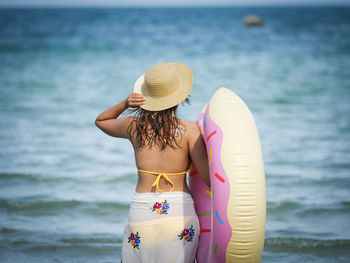 Rear view of woman wearing bikini carrying inflatable ring while standing at beach