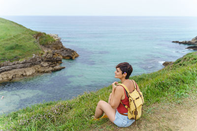 Rear view of woman sitting on cliff by sea against sky