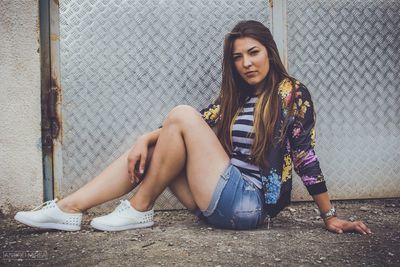 Full length portrait of young woman sitting against metallic door