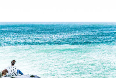 Rear view of woman sitting by seascape against clear sky