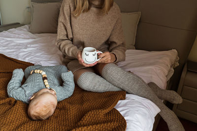 Low section of mother holding coffee cup sitting on bed with sleeping baby