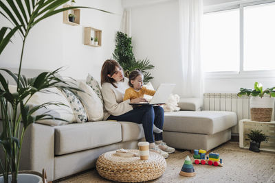 Mother sitting with daughter and using laptop in living room