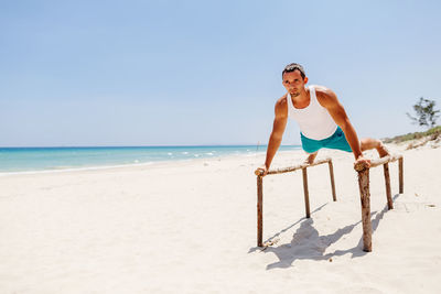 Man exercising on parallel bars at beach against blue sky