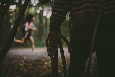 Midsection of man with rope standing against running woman in forest