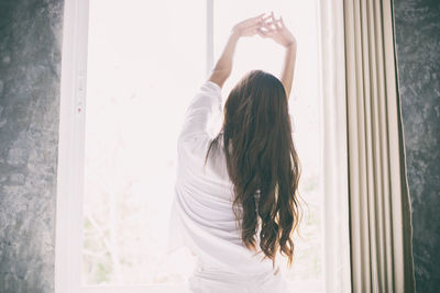 Rear view of woman stretching arms by window at home