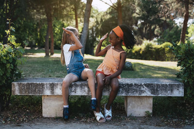 African american girl smiling and giving high five to best friend sitting together on bench in green park in sunny day