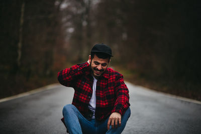 Carefree man in stylish casual wear sitting on asphalt road and looking down with closed eyes among autumn forest