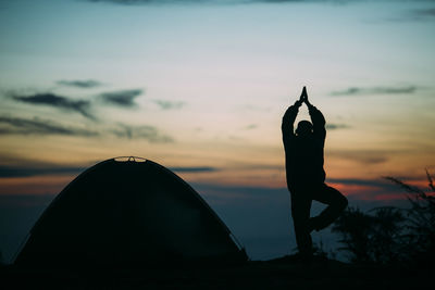 Silhouette man practicing tree pose by tent against sky