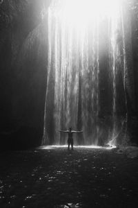 Rear view of man standing with arms outstretched by waterfall in forest