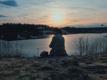 Rear view of woman sitting by lake against sky during sunset