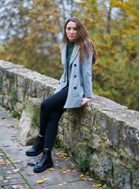 Portrait of a young caucasian woman with brown hair on a stone bridge with the green of the trees in the background. dressed in a gray jacket, green sweater, dark pants and military boots