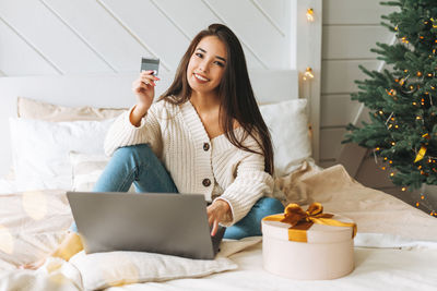 Young asian woman with gift box using laptop with credit card in room with christmas tree at home