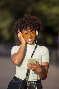 Young woman with afro hair and glasses smiling happy using smartphone and earphones in the city.