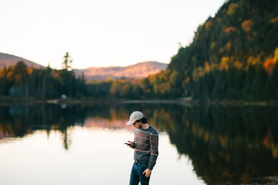 Young stylish male tourist in warm sweater and cap messaging on smartphone while standing on shore of picturesque calm lake reflecting lush colorful autumn trees during trip in canada