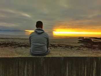 Rear view of man sitting on beach against sky during sunset