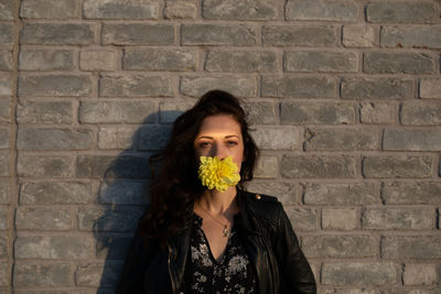 Portrait of woman with yellow flower in mouth against brick wall
