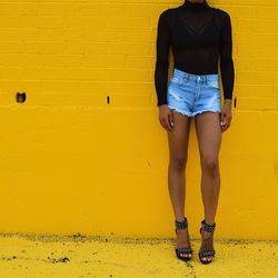 Low section of woman in hot pants leaning on yellow wall