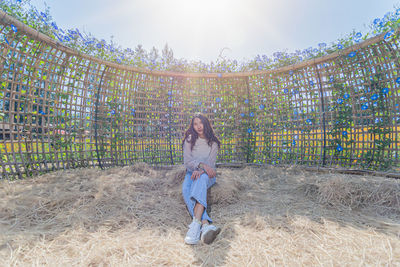 Portrait of young woman sitting on dry grass against wooden fence during sunny day