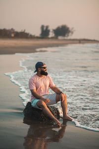 Side view of good looking bearded man sitting on beach during sunset with feet in the water