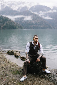 A serious lonely brutal hipster man in a wedding suit by the sea and mountains in nature outdoors