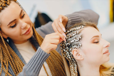 Young woman tying braids of female customer in salon