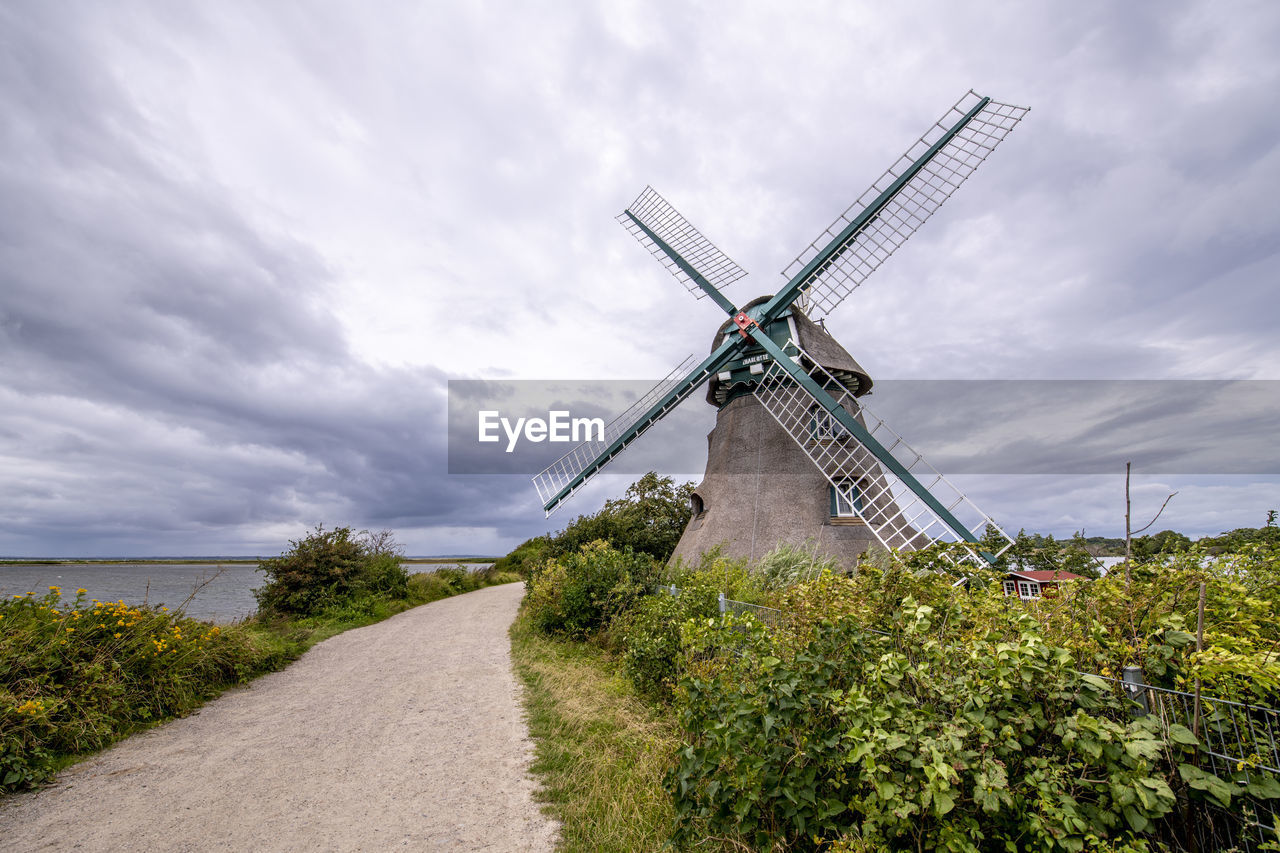 A classic windmill in the green, separated from the water by a path in front of a very cloudy sky.