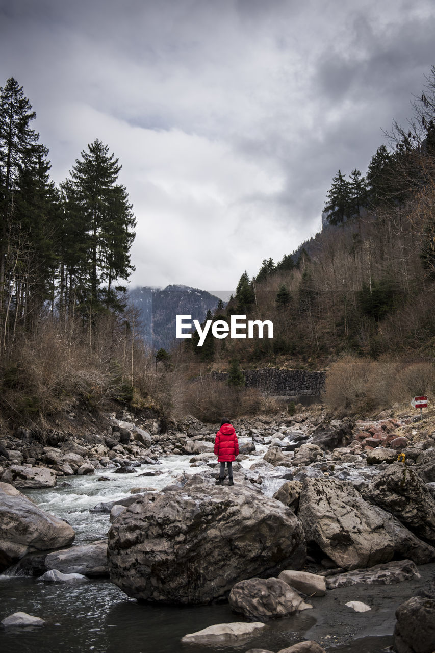 Girl standing on rock by stream flowing in mountains against sky