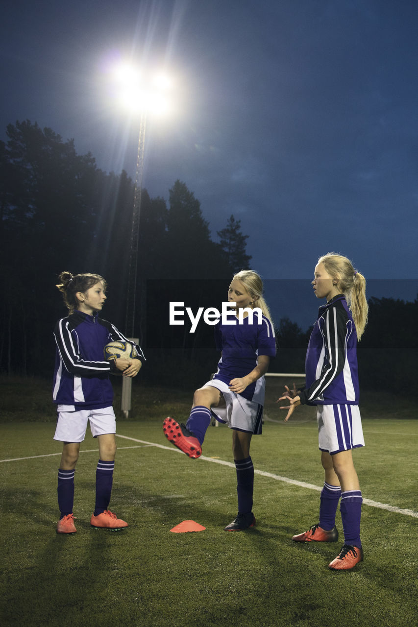 Girls talking while standing on soccer field against sky at night