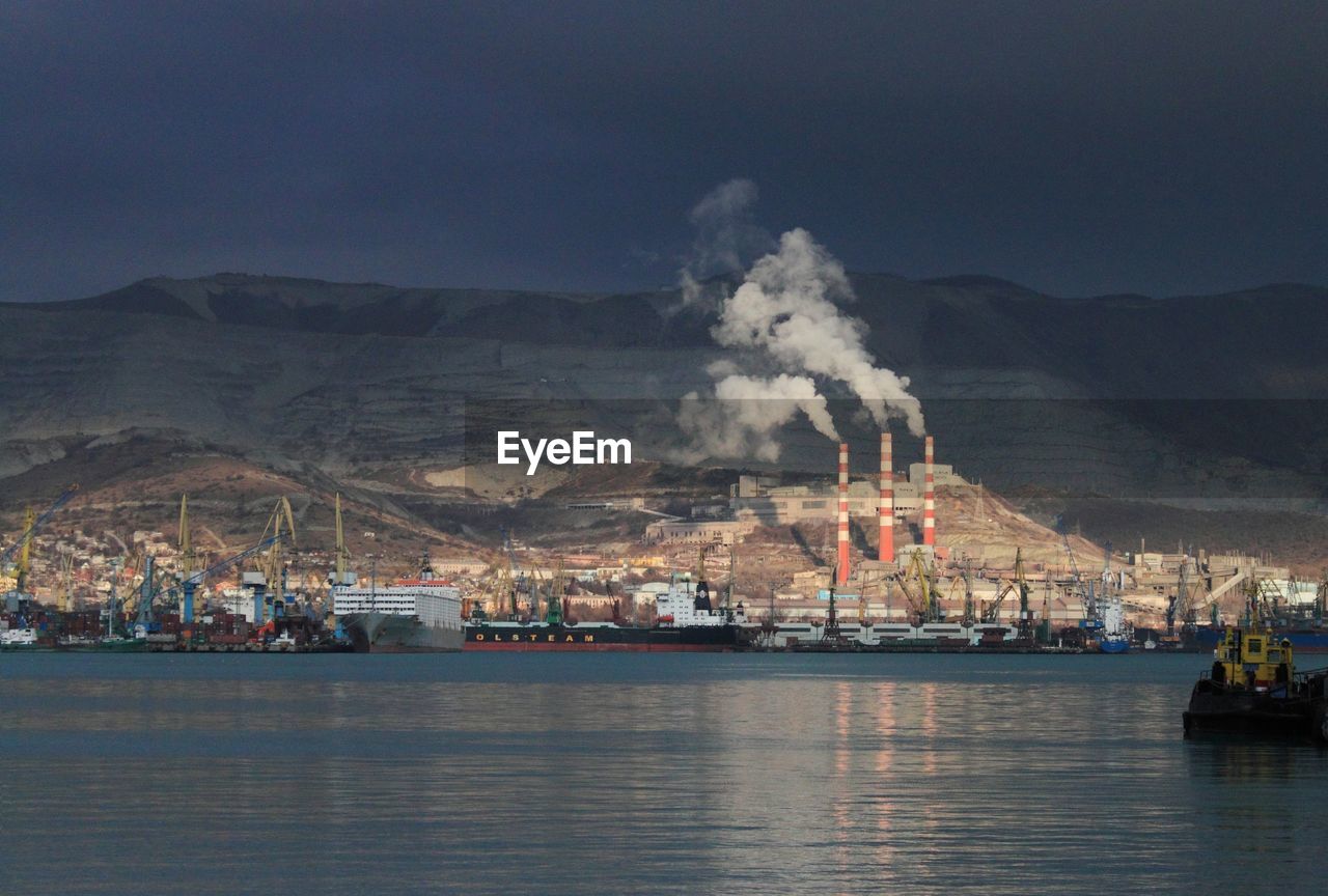 Commercial dock with ships and chimneys emitting smoke by mountain against sky