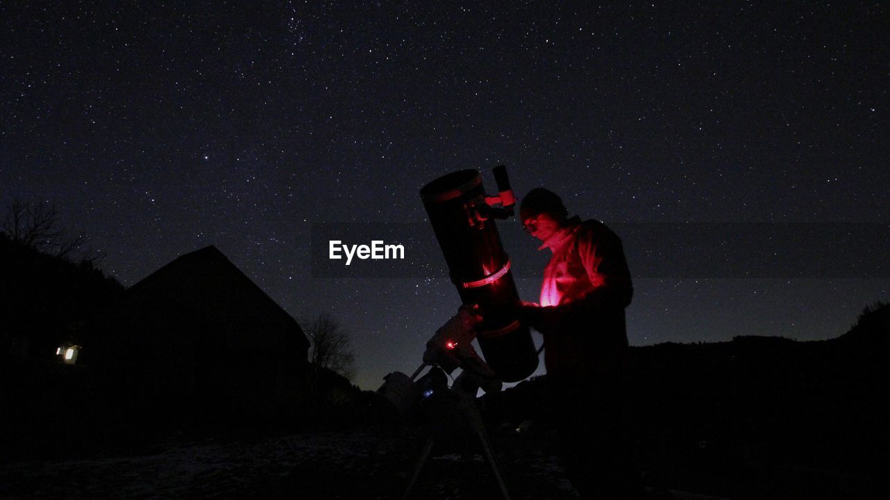 Man using mobile phone by telescope against sky at night