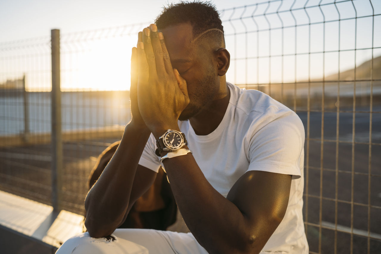 Thoughtful man with hands clasped sitting against fence