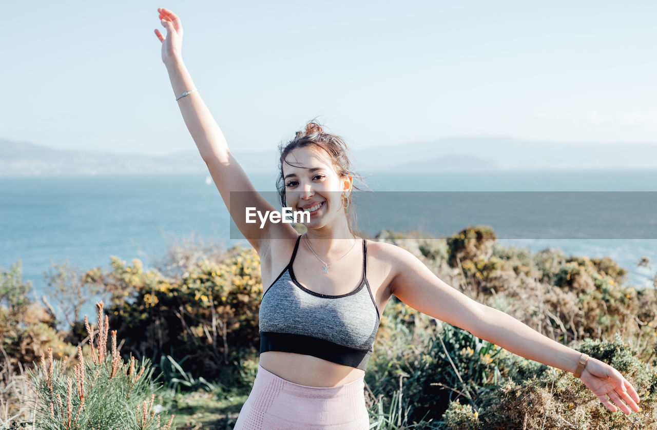 Side view of young woman with arms raised standing at beach against sky