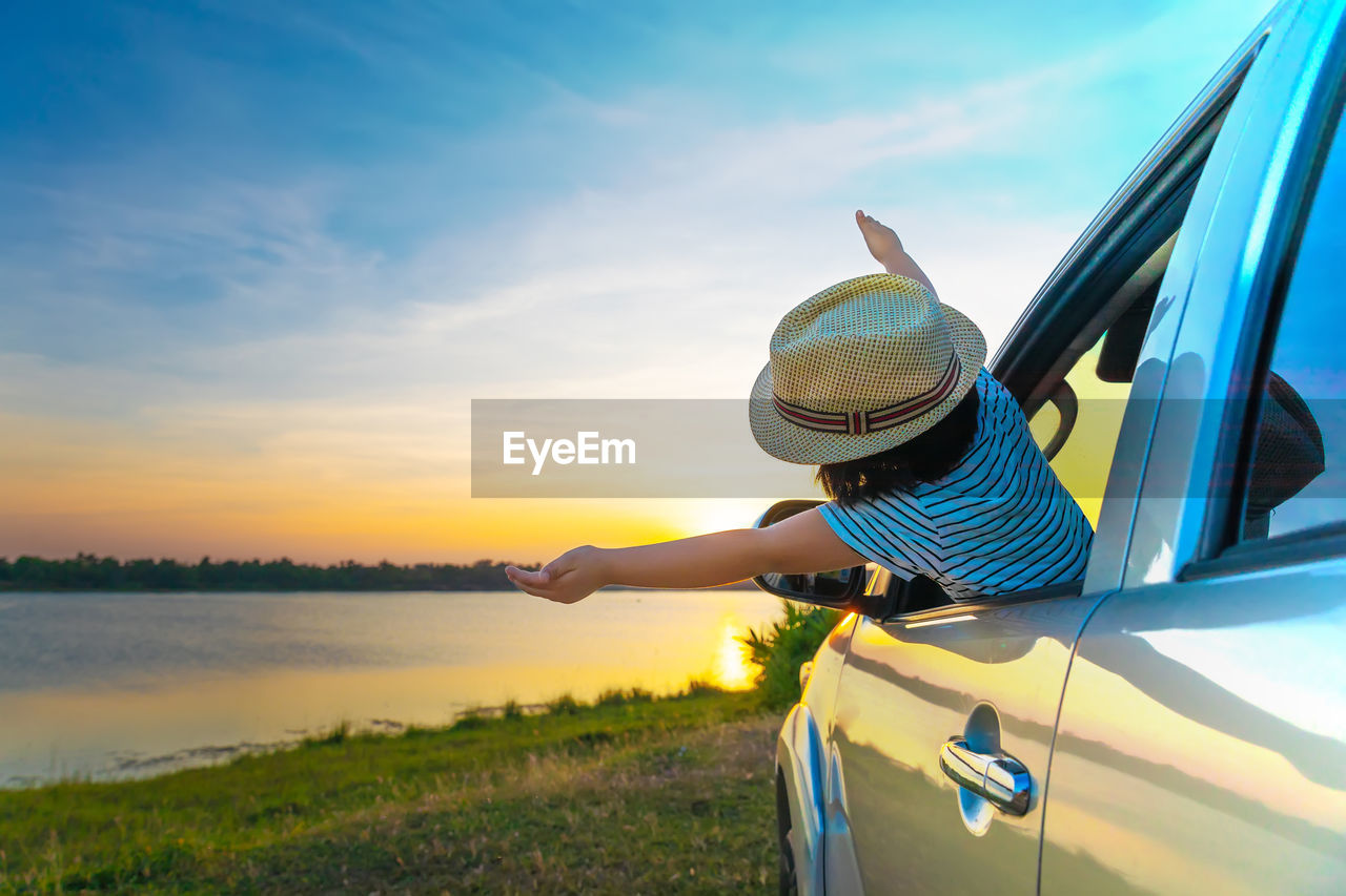 Rear view of girl wearing hat with arms raised peeking through car window against sky during sunset